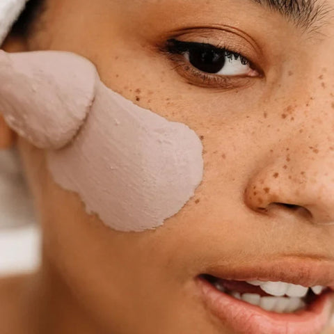 Close-up of a person applying a thick, pale pink cosmetic cream or mask onto their cheek with a spatula, showing freckled skin and part of their eye.